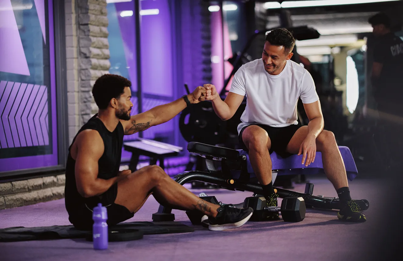 Two young men in a gym exchange a fist bump, celebrating fitness and camaraderie, with weights and workout equipment around them.