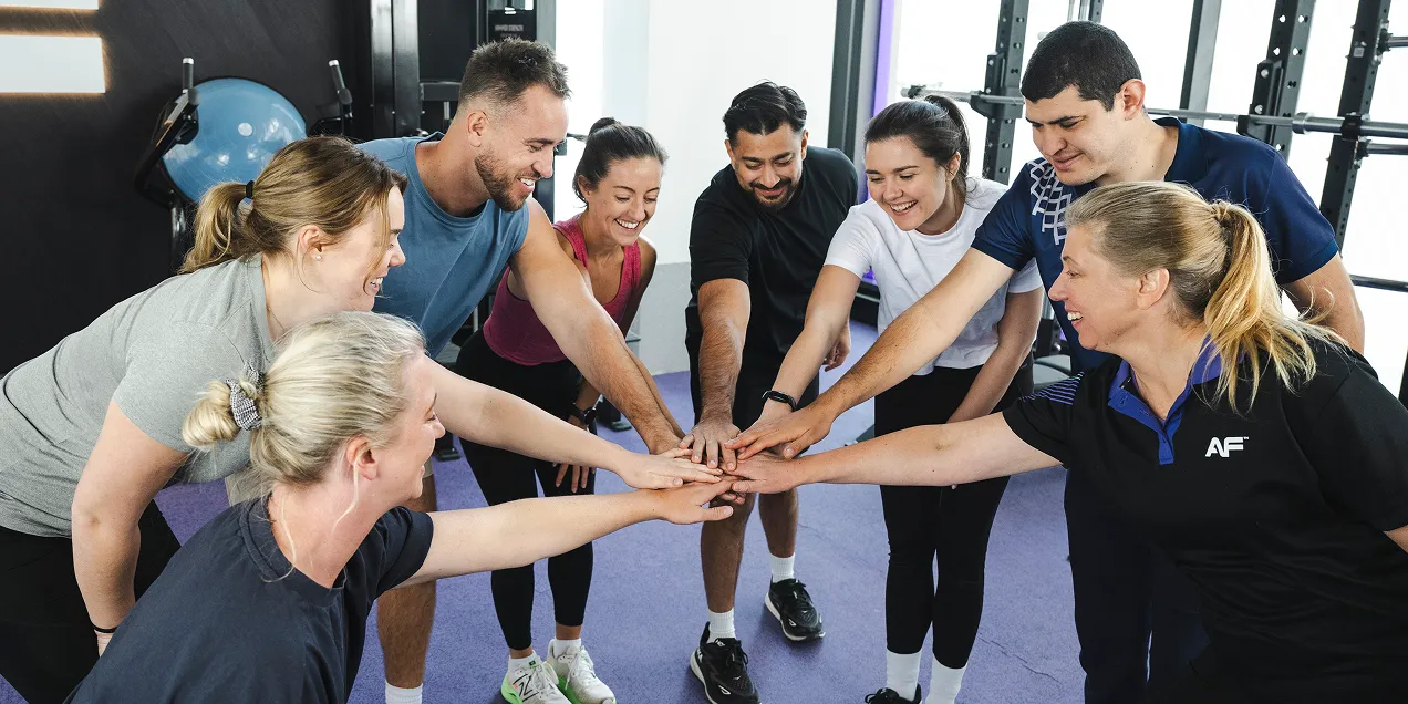 A group of diverse individuals in sportswear gather, placing their hands together in a show of teamwork and unity in a gym setting.