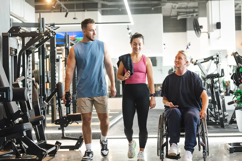 Three individuals walk through a modern gym, one in a wheelchair, promoting inclusivity in fitness. Exercise equipment is visible in the background.