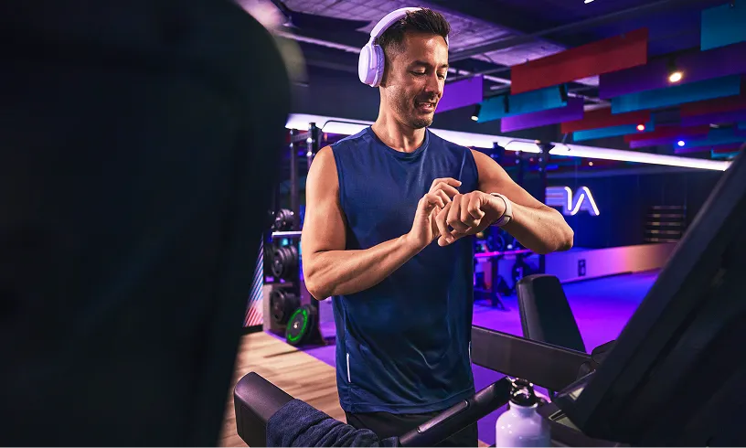 A person in a sleeveless shirt checks a smartwatch while exercising in a modern gym with vibrant lighting and equipment.