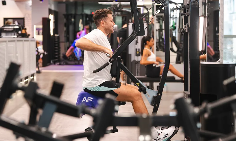 A man exercises on a cable machine in a modern gym, focusing on upper body strength, with another person working out nearby.