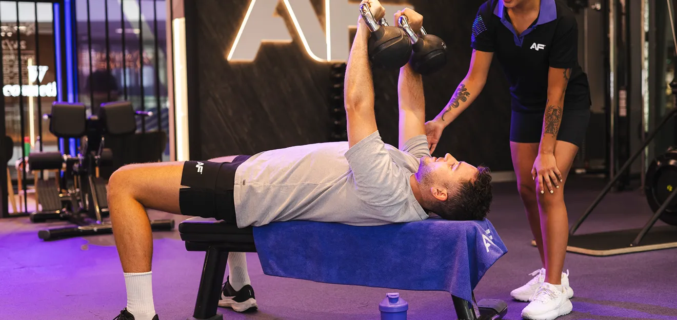 A person lies on a bench performing a kettlebell exercise, while a trainer assists beside them in a modern gym setting.