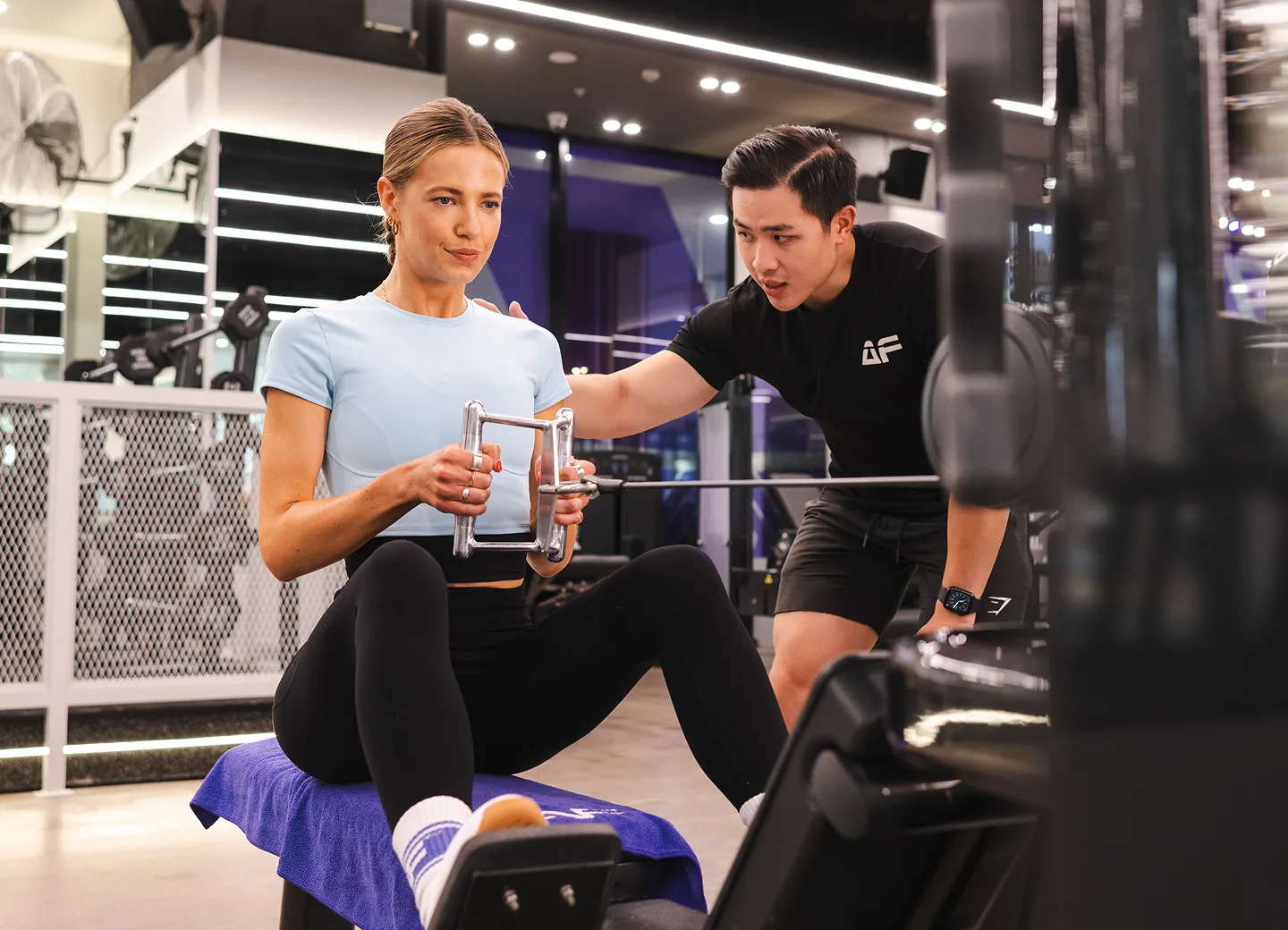 A woman in a light blue workout top is engaged in strength training with a trainer, using a resistance machine in a modern gym setting.
