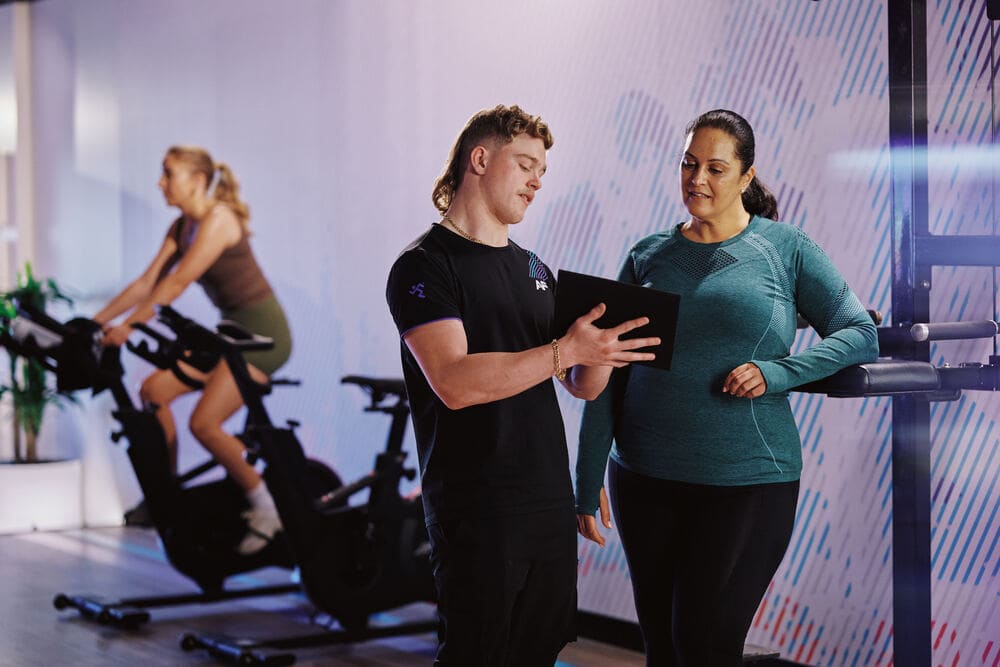 A personal trainer provides guidance to a client while another person exercises on a stationary bike in a modern gym environment.