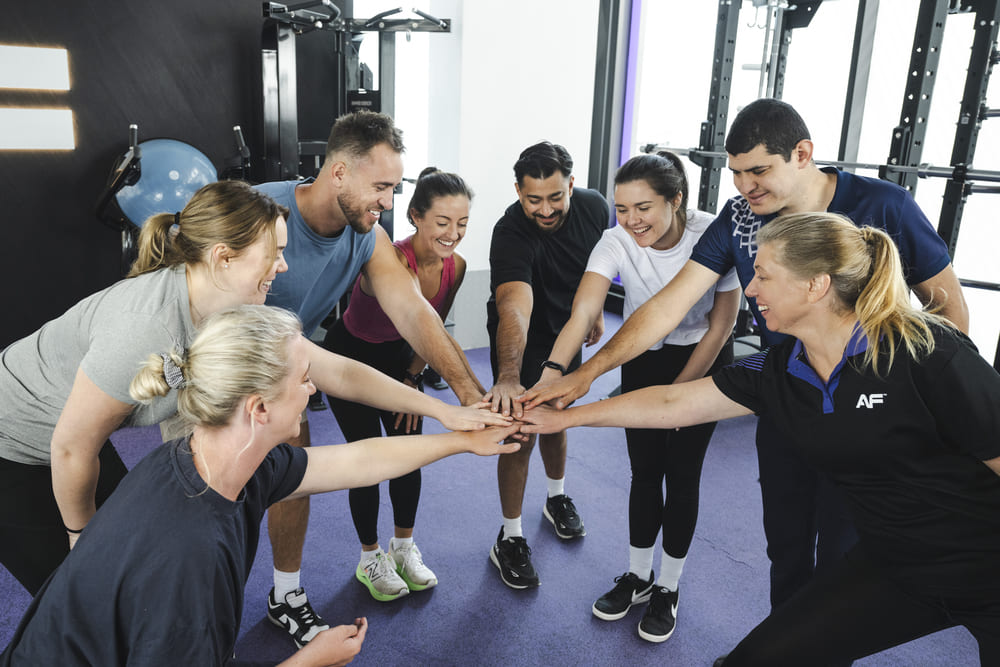 A group of individuals in workout attire gathers in a circle, placing their hands together in a show of teamwork and motivation.