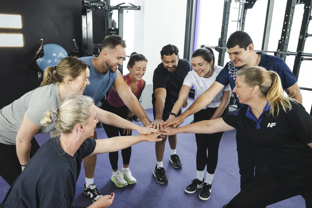 A group of individuals in workout attire gathers in a circle, placing their hands together in a show of teamwork and motivation.