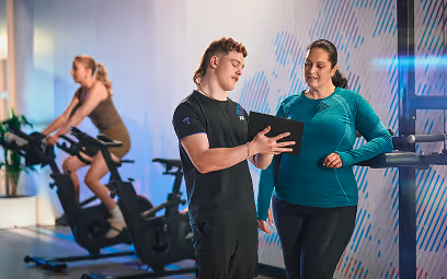 A fitness coach shows a workout plan on a tablet to a gym member at Anytime Fitness, with other members exercising on stationary bikes in the background.