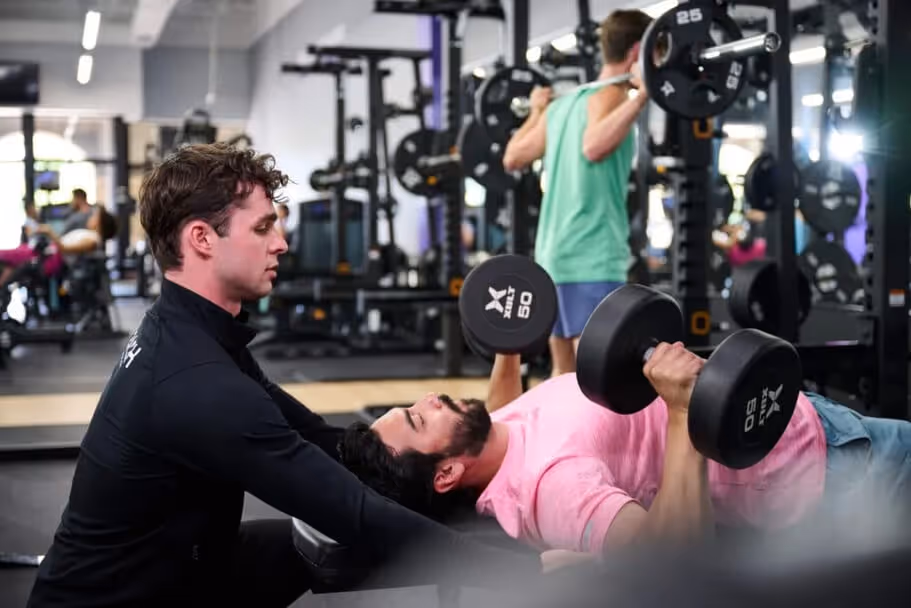 A personal trainer assists a client with dumbbell bench presses in a modern gym, while others work out in the background.