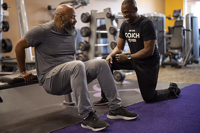 A man performs dips on a bench while a fitness coach kneels beside him, offering guidance in a gym setting filled with equipment.