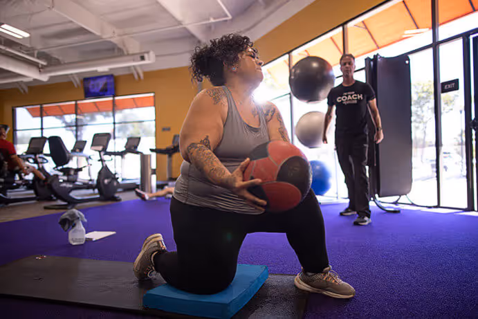 A person kneels on a mat, holding a medicine ball, while a trainer observes in a brightly lit gym with exercise equipment in the background.