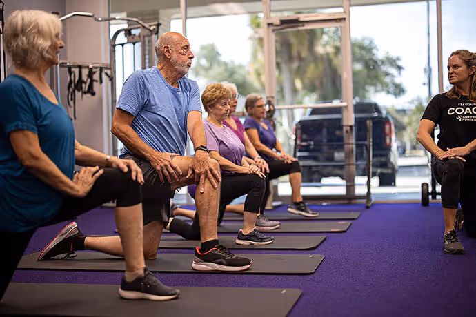 A group of seniors engages in a stretching exercise on mats in a bright fitness studio, led by a coach.