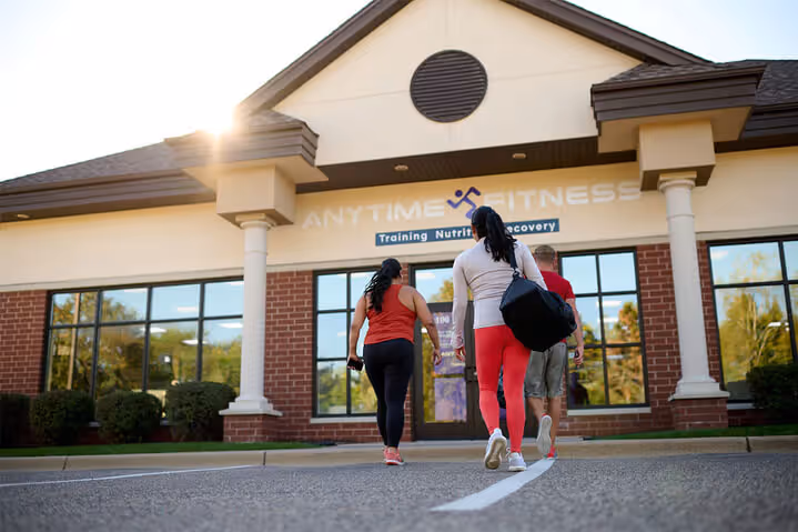 Three people walking towards an Anytime Fitness gym, wearing workout clothes and carrying bags, illuminated by soft sunlight.