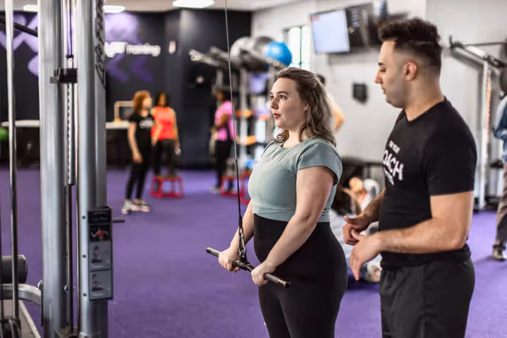 A fitness coach assists a woman with an exercise at a gym, while others work out in the background on purple carpeted flooring.