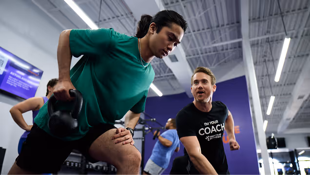 A man in a green shirt performs a single-arm kettlebell row exercise while a fitness coach, wearing a "I'm Your Coach" shirt, instructs him in a group gym setting.