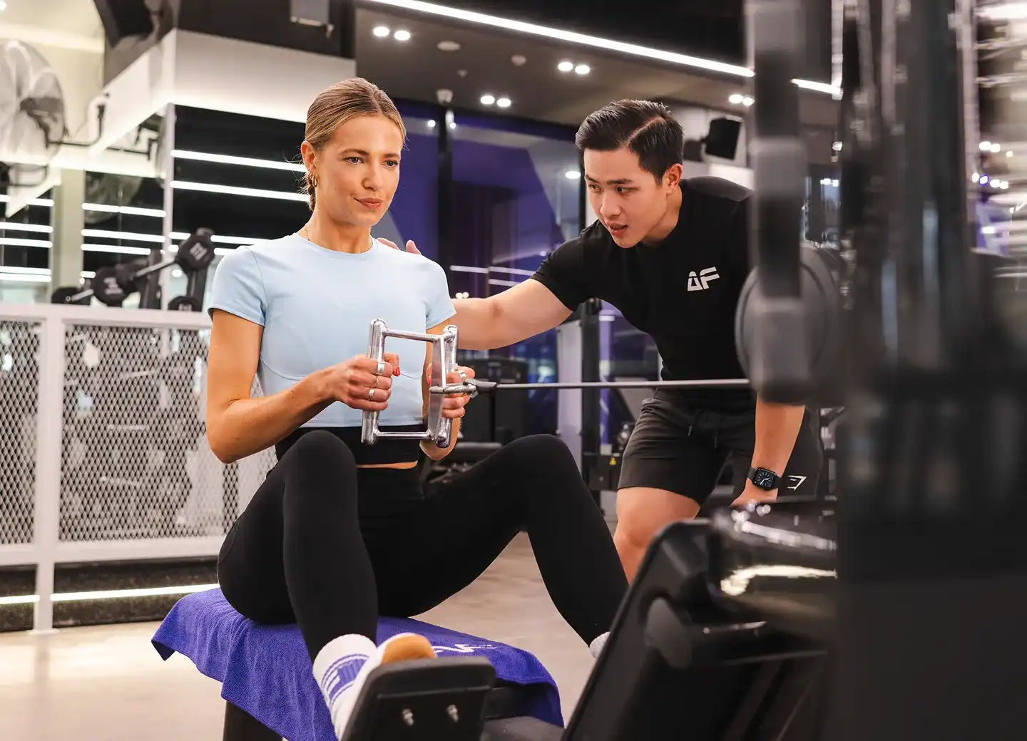 A woman in a light blue workout top is engaged in strength training with a trainer, using a resistance machine in a modern gym setting.