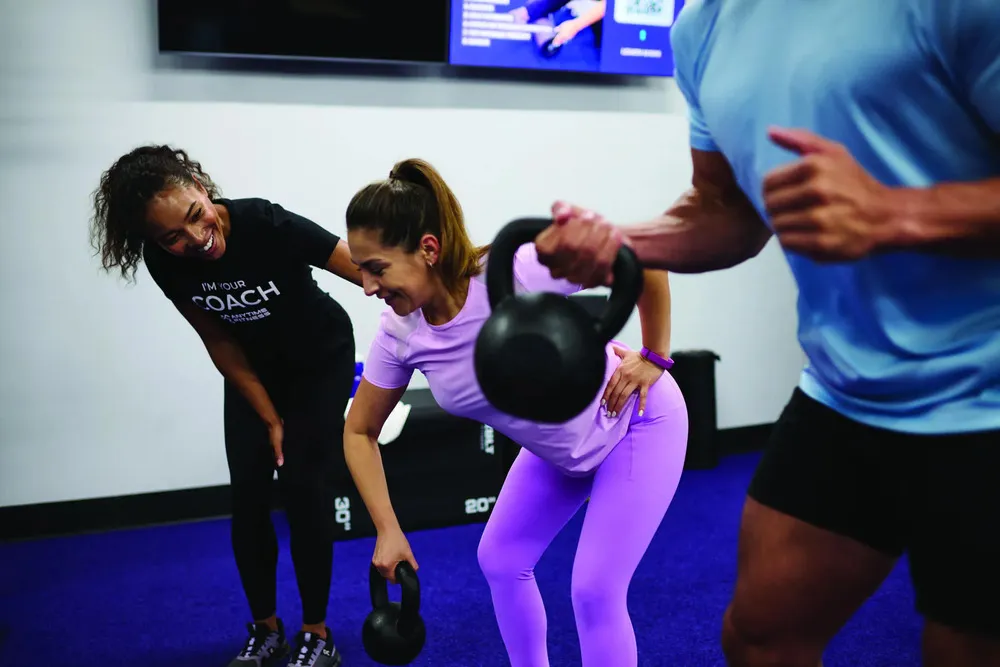 A person in workout attire performs an exercise with a kettlebell, while another individual observes and coaches nearby in a gym setting.