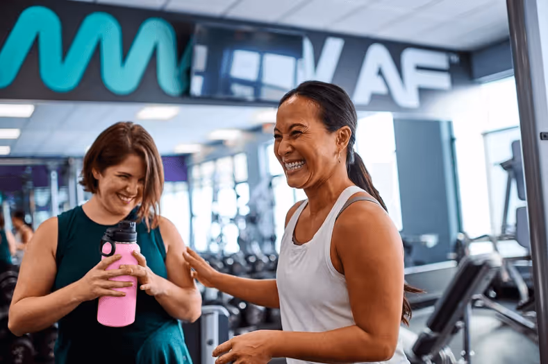 Two women smiling and talking inside a gym near cardio equipment