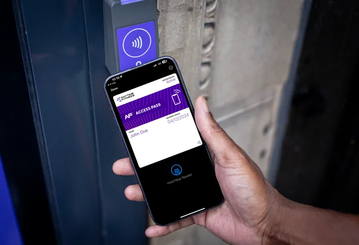 A hand holds a smartphone displaying a digital access pass while near a contactless reader by a door.