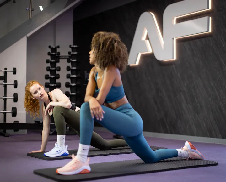 Two individuals practice stretching exercises on yoga mats in a modern gym with exercise equipment in the background.