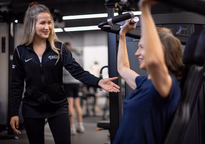 A female personal trainer in a black Anytime Fitness jacket smiling while assisting a client on a lat pulldown machine.
