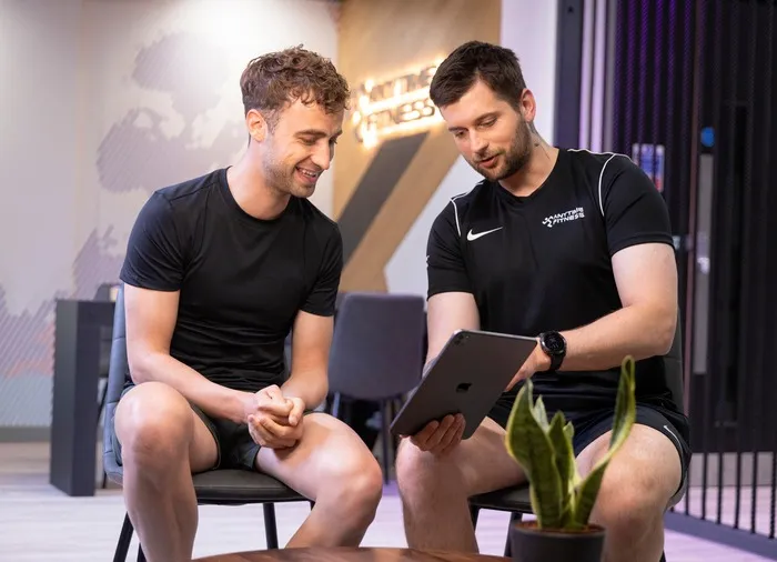 Two men in athletic wear sit in a modern fitness studio, discussing a tablet while a green plant is visible on the table.