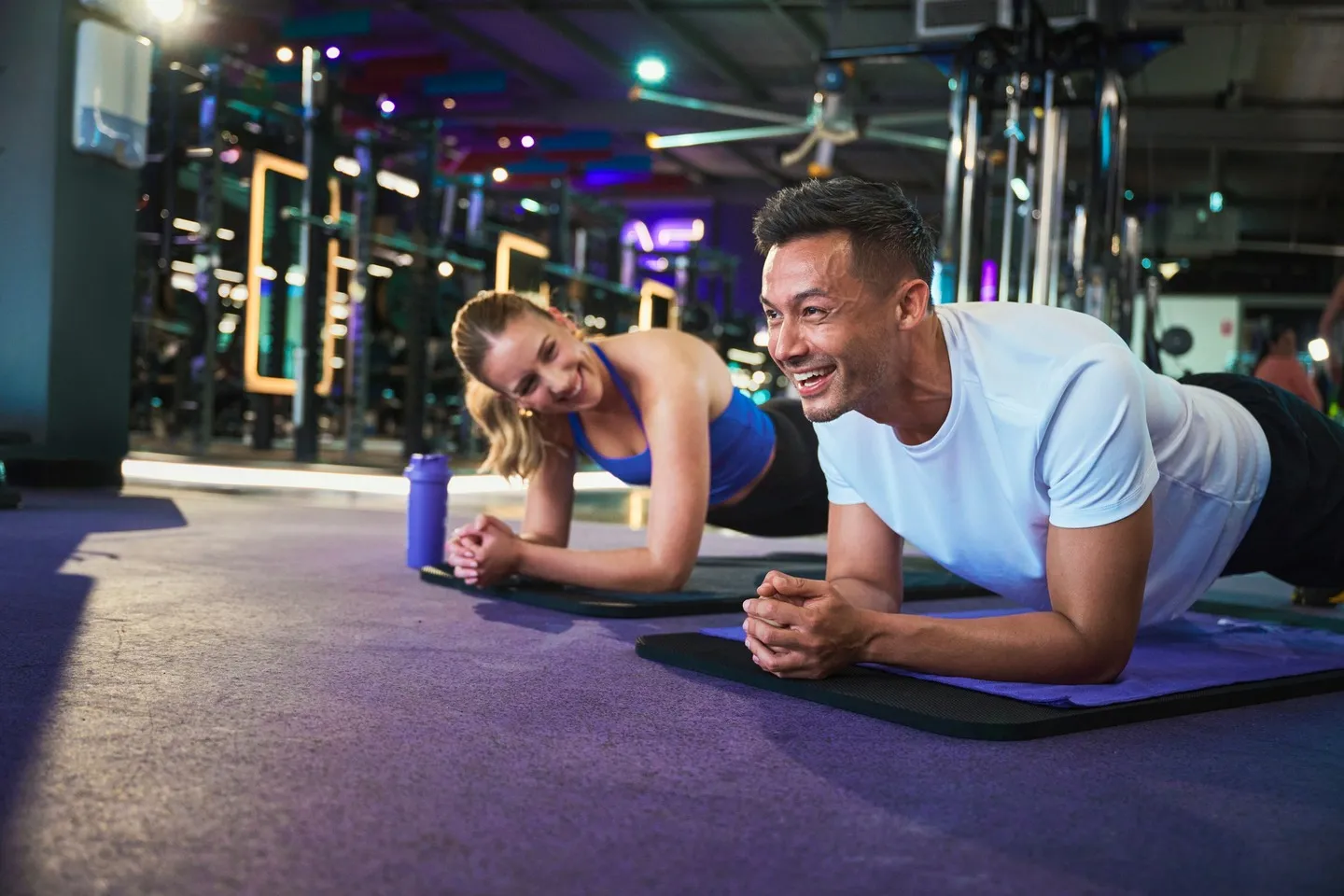 Two people smiling while holding a forearm plank on exercise mats in a modern gym, with strength equipment in the background and a purple water bottle beside them.