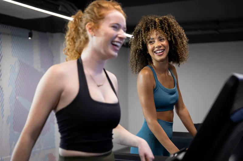 Two women in athletic wear using treadmills in a modern gym with dark ceiling and lighted panels.
