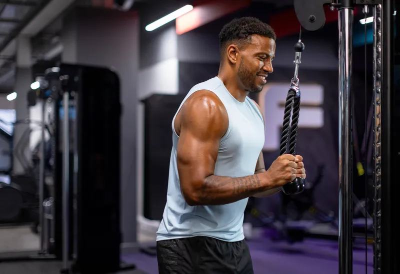 Man with muscular arms in a sleeveless shirt using a cable machine for triceps exercise in a gym.