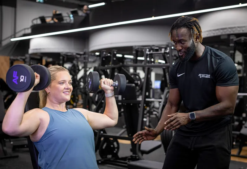 Woman lifting dumbbells during a shoulder press exercise while a male trainer provides guidance in a gym setting.