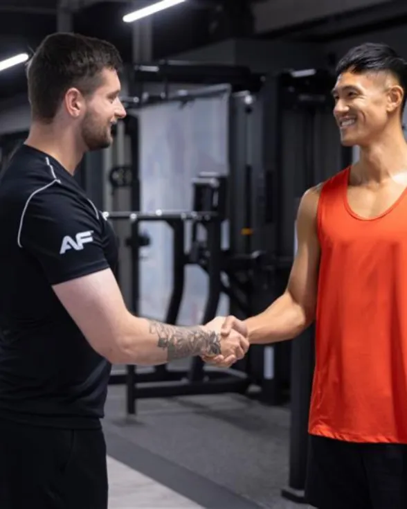 Two men shaking hands inside a gym, one in a black shirt and the other in an orange sleeveless top.