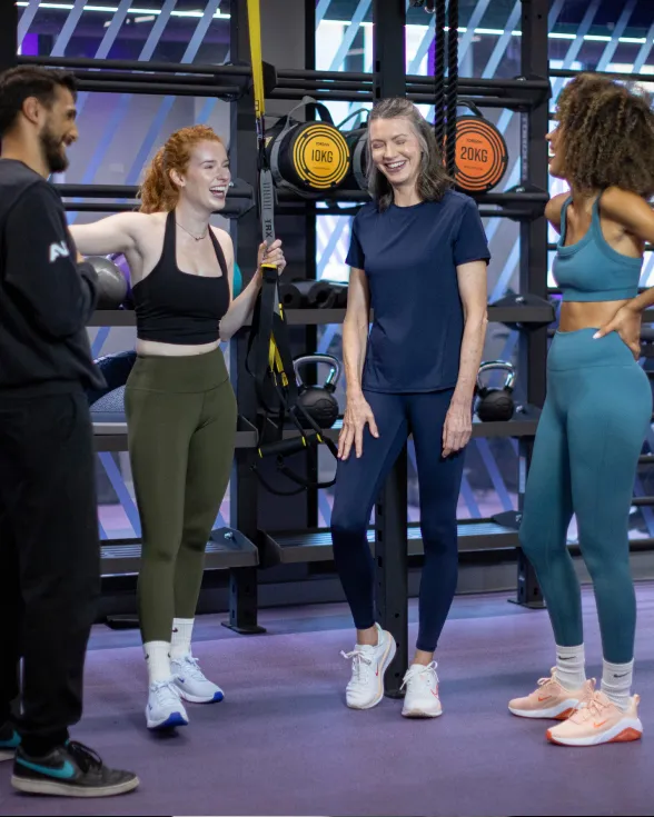 Group of four people in athletic wear standing and talking in a gym with weights and exercise equipment in the background.