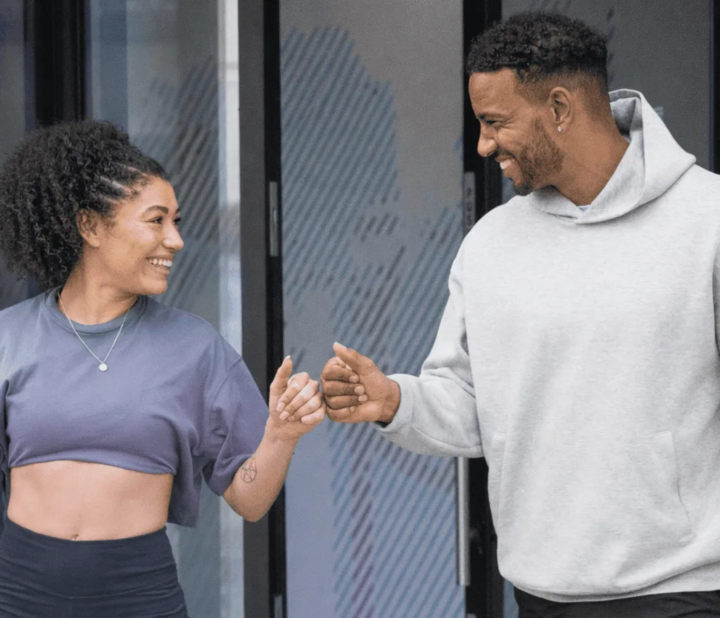 Two people outdoors fist bumping each other, one wearing a cropped t-shirt and the other a light gray hoodie.