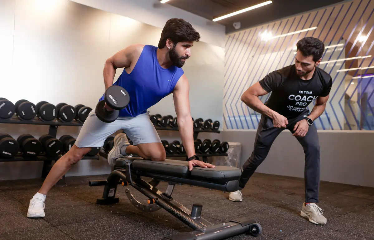 A personal trainer guides a client performing a one-arm dumbbell row on a bench in a modern Anytime Fitness gym, with rows of dumbbells in the background.