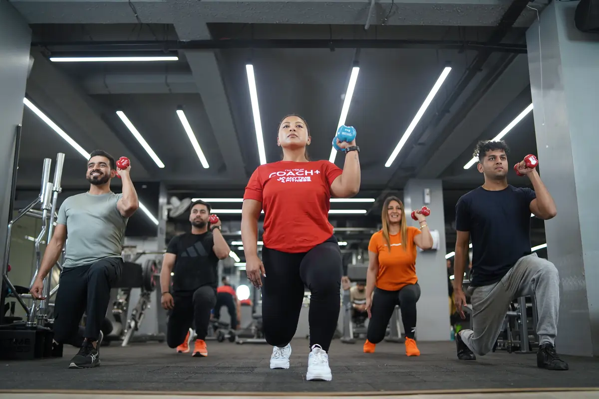 A group of members led by a trainer perform dumbbell lunges in a brightly lit Anytime Fitness gym, showcasing a motivating and energetic group workout environment.