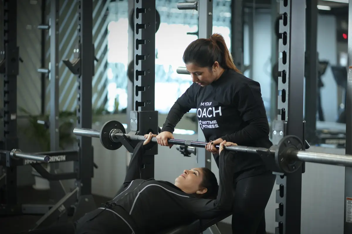 A personal trainer assists a member with a barbell bench press in an Anytime Fitness gym, ensuring proper form and safety during a strength training session.