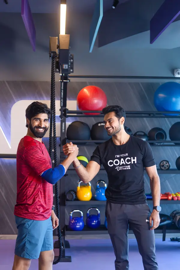 Two men in a modern gym smiling and clasping hands in a friendly handshake, with colorful kettlebells and exercise balls visible on racks behind them.