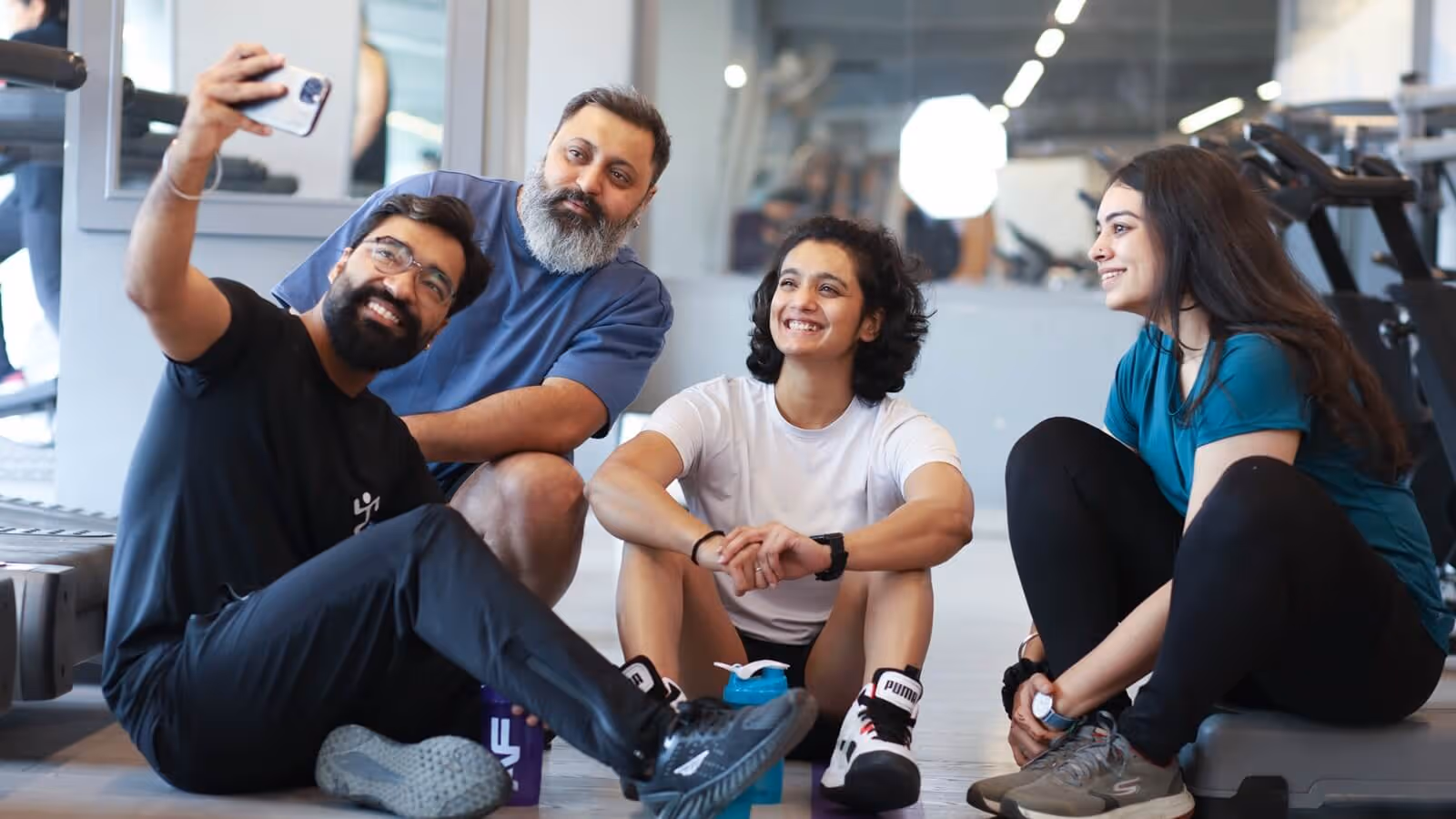A diverse group of coworkers smiling and taking a group selfie while sitting together on the gym floor.