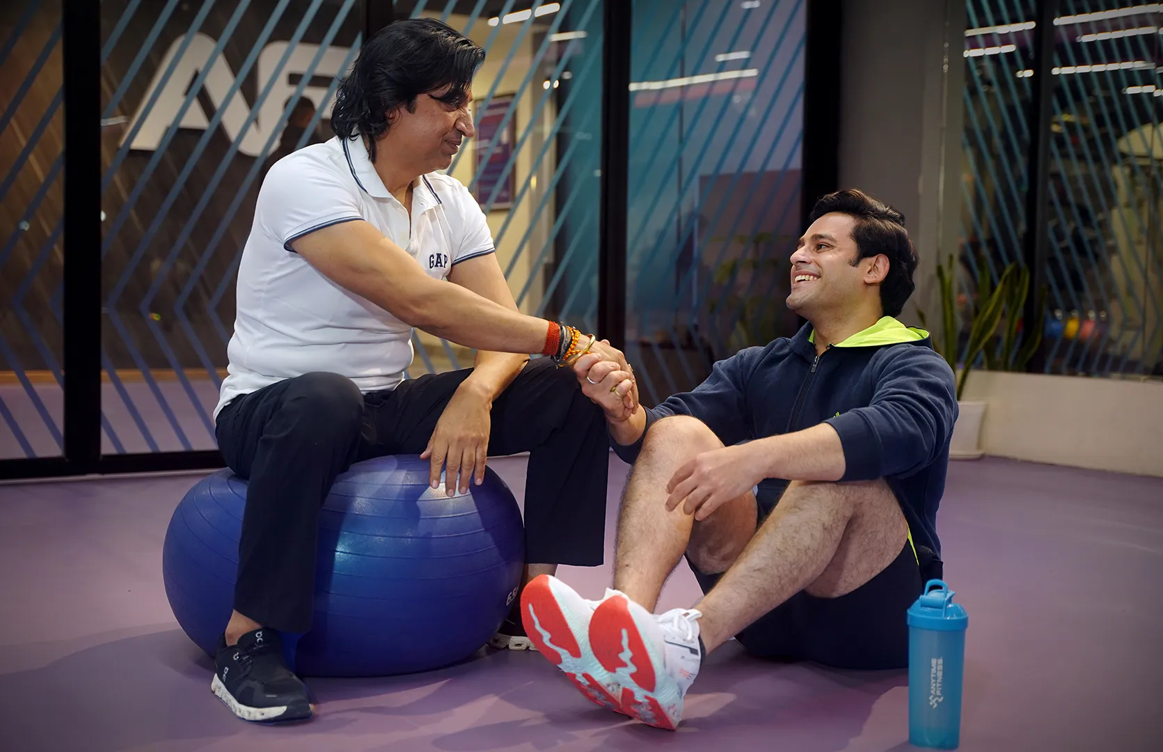 A vibrant, high-energy photo of a man and woman during a personal training session in a modern gym. The man, in a dark blue t-shirt, is performing a bicep curl with a dumbbell while a female trainer in a black sports bra and leggings stands beside him, smiling and providing motivation. The background is softly blurred, focusing the viewer on the supportive and personalized coaching experience at Anytime Fitness.