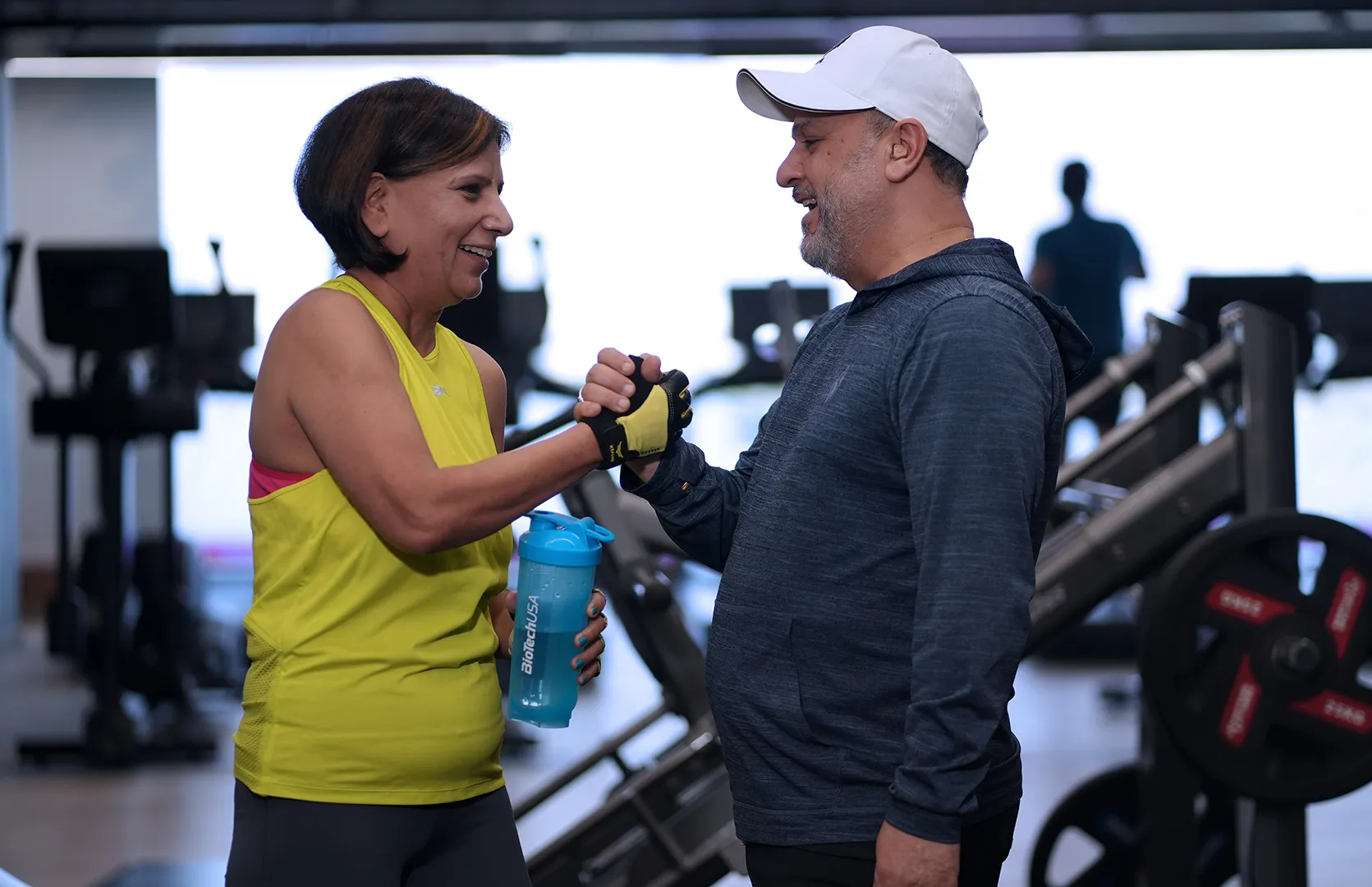A bright and welcoming image of a fitness professional and a client engaged in a health consultation. A man with a beard and a blue t-shirt stands next to a woman in a black athletic top; both are looking at a tablet together and smiling. The setting is a clean, well-lit Anytime Fitness gym, emphasizing a friendly, no-pressure environment for members to discuss their fitness goals.