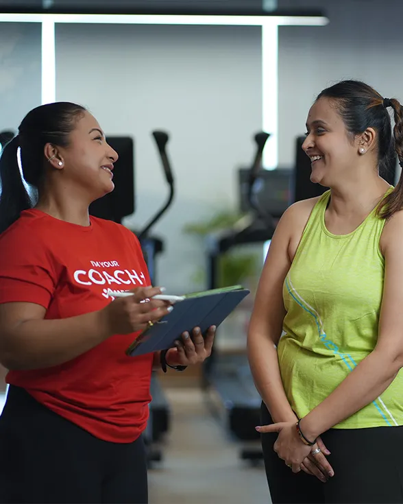 A gym coach in a red shirt holding a tablet talks and smiles with a woman wearing a green tank top in a fitness center.