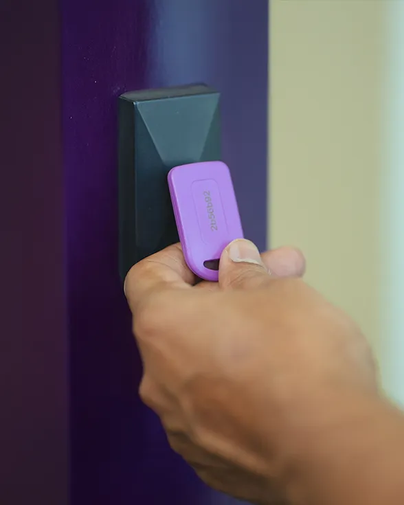 Hand holding a purple key fob near a black RFID reader attached to a purple door frame.
