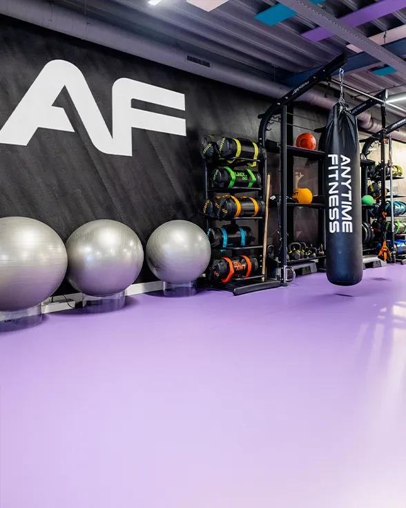 Modern gym interior with stability balls, colorful weighted bags, kettlebells, and a black punching bag with Anytime Fitness logo on a purple floor.