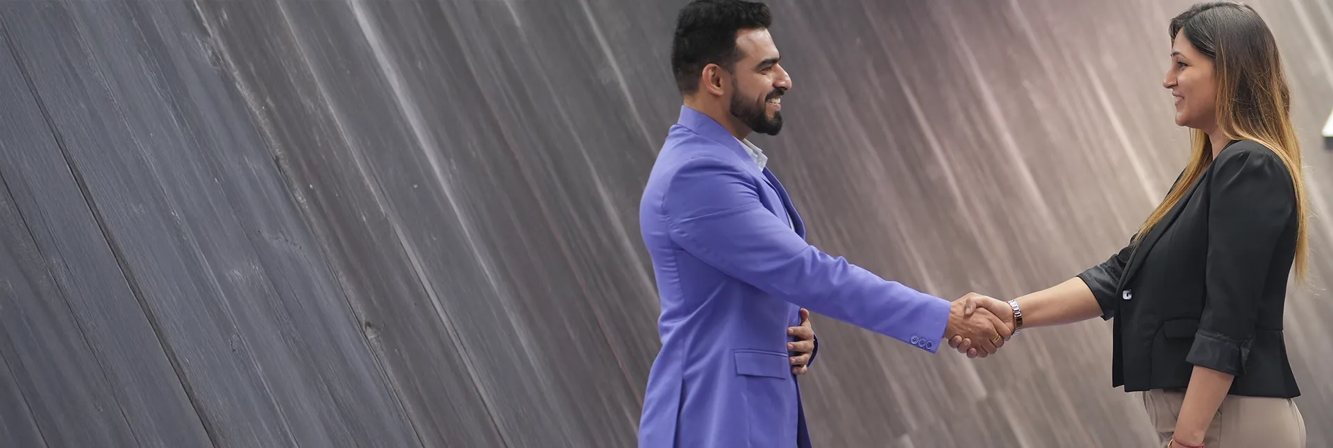 Man in blue blazer and woman in black blazer shaking hands and smiling indoors.