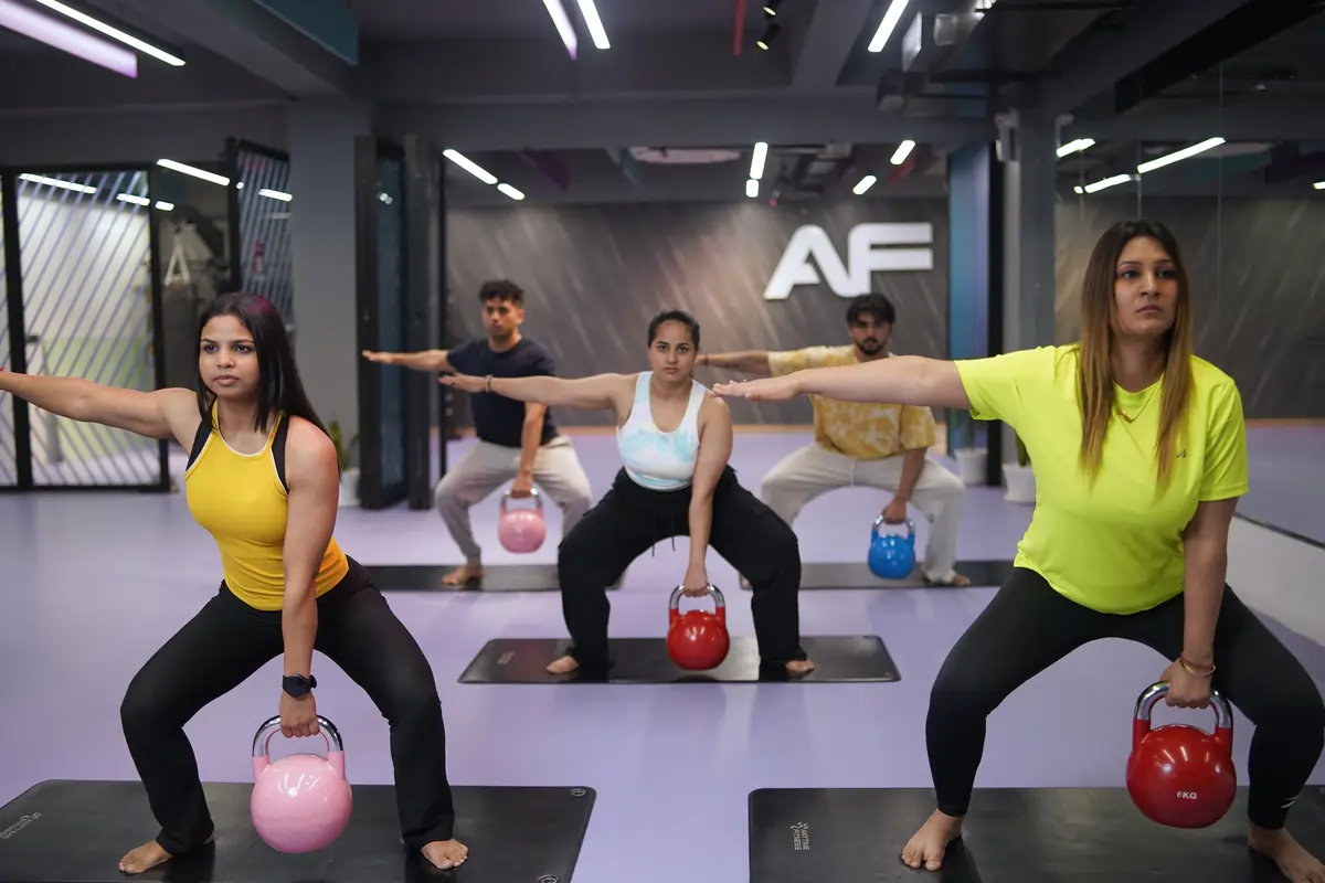 Group workout class with five people holding kettlebells in a squat position with one arm extended for balance in a gym.