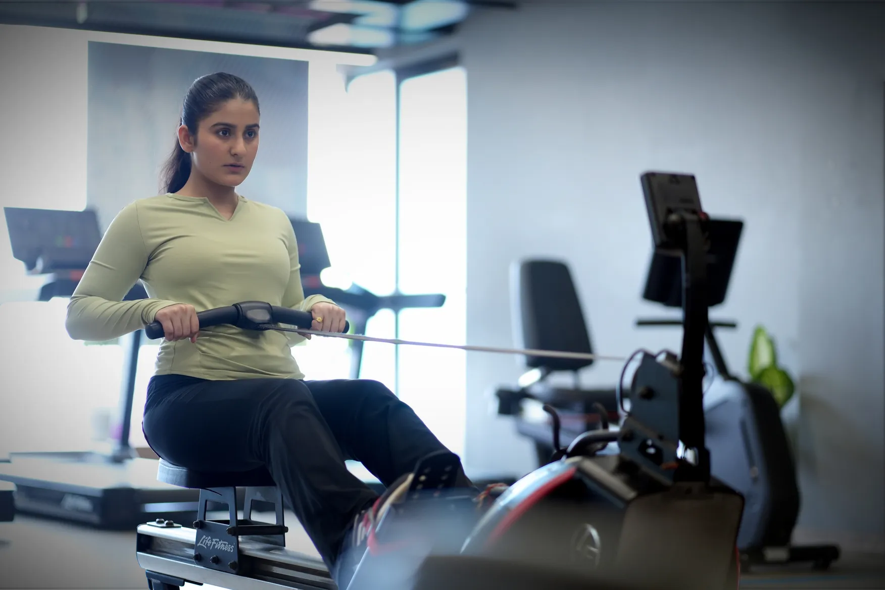 Woman wearing a green long-sleeve top using a rowing machine in a gym setting.