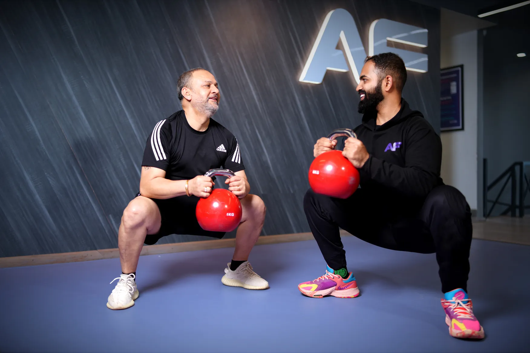 Two men squatting with red 6kg kettlebells inside a gym with AF logo on the wall.