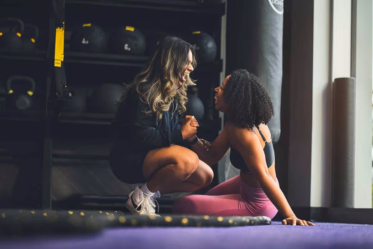 Two women smiling and holding hands in a gym, one kneeling and the other sitting on the floor.