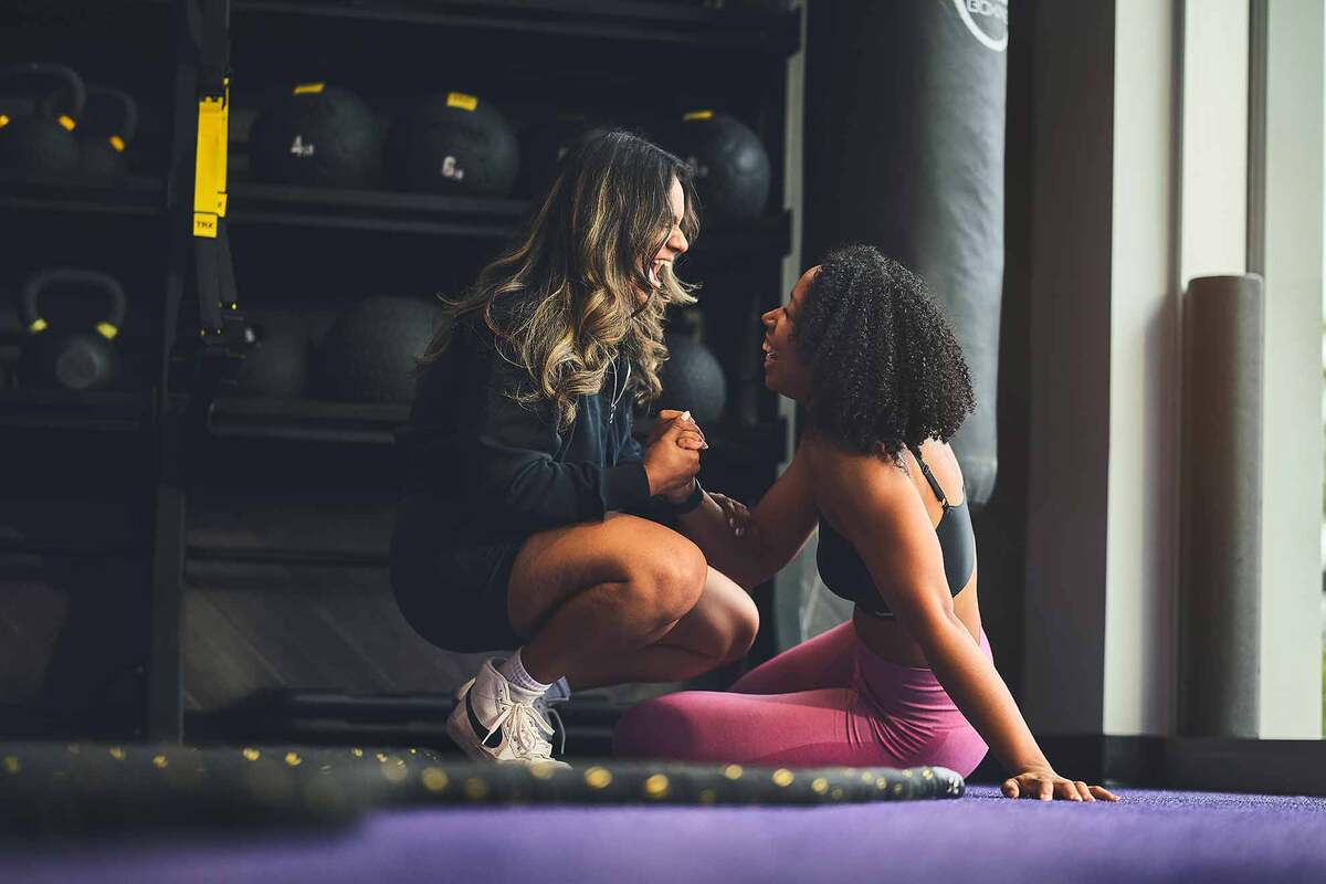 Two women in workout clothes supporting each other in a gym with kettlebells and a punching bag in the background.