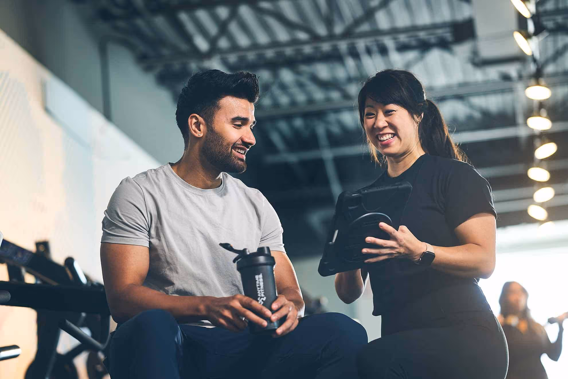 Woman showing a tablet to a man holding a water bottle at a gym, both smiling.
