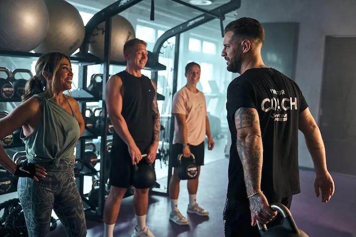 Ein Fitness-Coach von Anytime Fitness im Gespräch mit drei lächelnden Mitgliedern während einer Trainingseinheit mit Kettlebells im Studio.